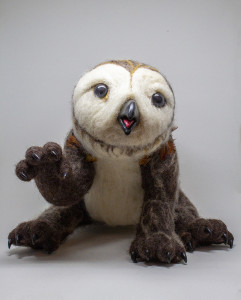 A felted, 3D owlbear stares at the camera. Its legs are dark brown, and one forelimb stretches towards the cameera. It has a white belly and face, which is owl-shaped complete with beak.
