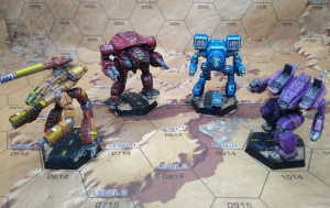 Four hand-painted miniatures of battletech lancers stand on a hex grid map. Each has been painted the colors of a Desert Bus shift. From left to right, they are gold, crimson, sky blue, and dark blue.