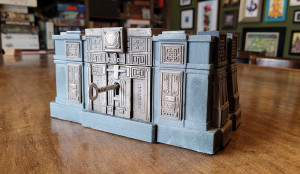 A bluish gray box with gleaming silver panels. The panels have each been intricately carved with runes and symbols. There is a key inserted into the box.