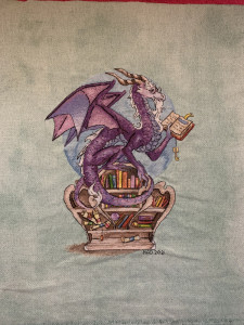 An intricately stitched purple dragon perches on top of a bookshelf shaped like an armchair. The dragon is wearing spectacles and reading from a book with a threaded key hanging from it as a bookmark.