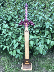 Wooden replica of the Master Sword, complete with tiny triforce, displayed in front of a leafy green bush.