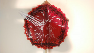 A red acrylic design of a spike-edged circle laid onto a pentagonal wooden base. The edges of the circle are decorated with black lines, and the center image features the silhouette of a person with arms spread and white blast of power shooting from in front of their chest.