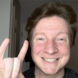 A headshot of a person who uses he/him pronouns. Andy has short reddish hair and smiles broadly at the camera, his right hand lifted in the ASL symbol of "I love you."