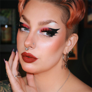 A glamorous headshot of a person who uses she/her pronouns. Her orange-dyed hair is cropped short on top of her head and features a side shave. Her face glows with lush red lipstick and deep pink eyeshadow that fades into a perfectly edged black wing shape at the corner of her eye.