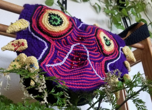 A crocheted messenger bag with a rendition of Majora's Mask. The overall bag is blue, with big yellow eyes and a pattern of pink stripes.