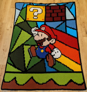 A crocheted blanket that shows Paper Mario about to hit a question mark block. Mario is trailing a rainbow behind him as he moved, and the green grass and blue sky around him are broken into triangular bits, like folded paper.