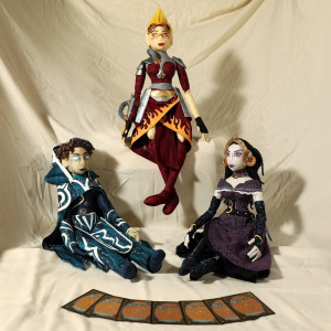 Three cloth dolls are arranged in front of a white sheet. From left to right, they are: Jace Beleren (blue), Chandra Nalaar (red), and Liliana Vess (purple).