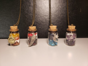 A lineup of four small glass bottles stoppered with corks. Each bottle has a different color of gemstones inside, starting from the left: yellow, red, blue, and purple. They also have metal emblems for each Desert Bus shift logo.