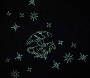 A close-up of a black blanket, pictured at night. The outline of the griffin, surrounded by stars, is glowing faintly green in the dark.