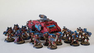 A collection of 14 Primaris minis, painted in Desert Bus colors, standing in front of a red transport bus with the Desert Bus logo on its side.