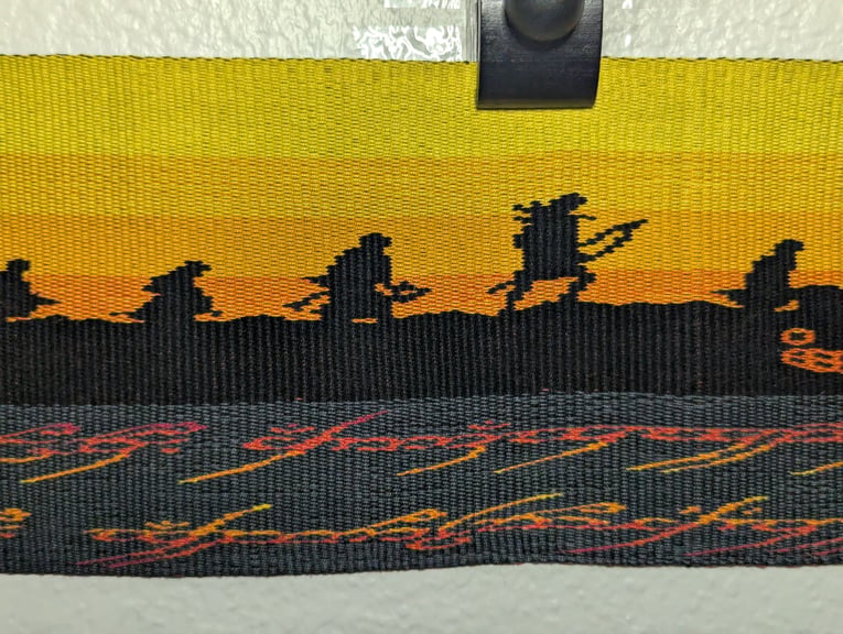 Fourth of six close ups of a tapestry depicting Pippin, Merry, Gimli, Legolas, and Frodo above part of the One Ring’s inscription.