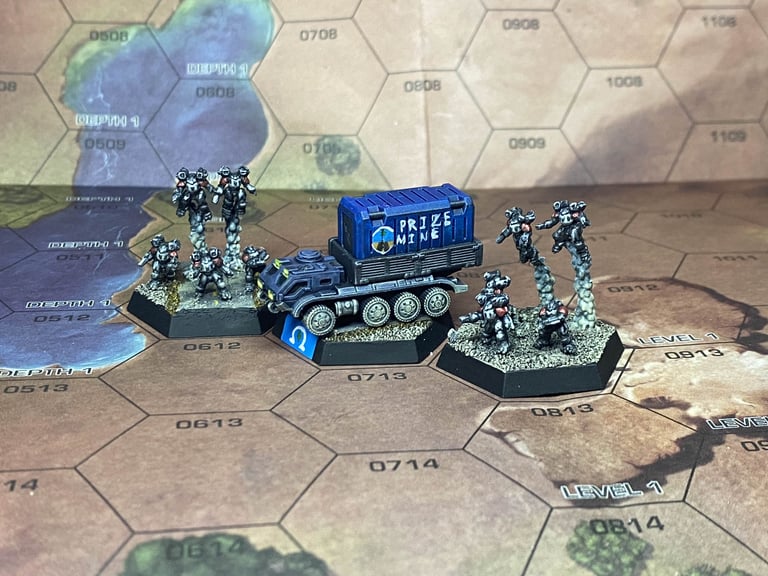 A heavy truck with a blue shipping container with "PRIZE MINE" written on the side with 2 groups of Elemental Battle Armor on ether side on a desert map
