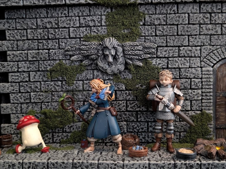 A close-up of several characters in the diorama. Marcille excitedly prepares to blast a walking mushroom into oblivion, while Laios sheathes his sword beneath a stone relief of a winged lion. Various bowls and food items litter the floor.