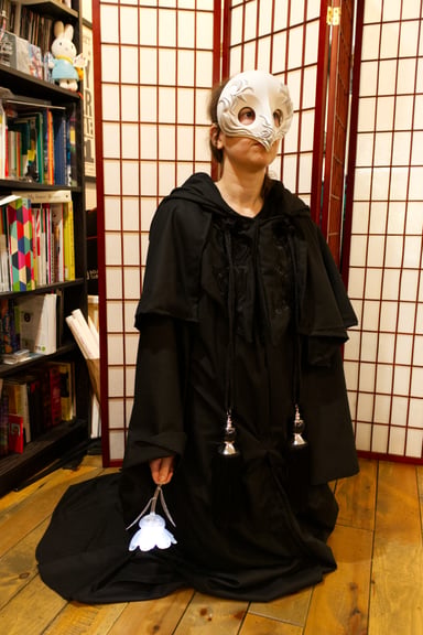 A full body shot of the robe and mask, including large floor length black robes with long robe cords decorated with metal silver, front lapel embroidery, a cowl, and an oversized hood. Note that the mask in this photo is just for the photograph, the mask in this Craft a Long is signed and should not be worn on the face to prevent it from rubbing off.