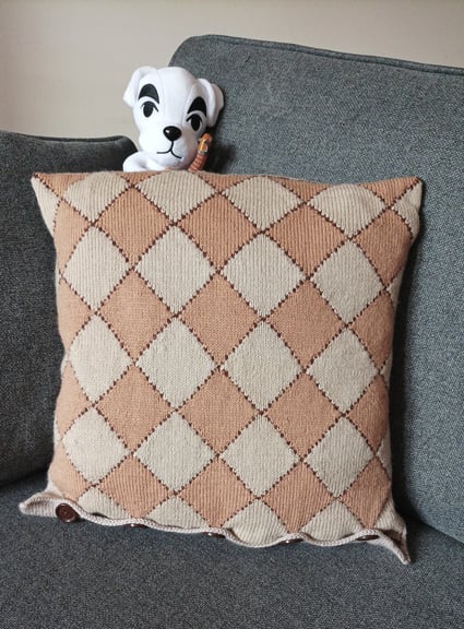 A photo of the back of the Blathers cushion, featuring a pattern of tan and cream diamonds, posed on a grey couch, with the head of a K.K. Slider plush poking out from behind it.
