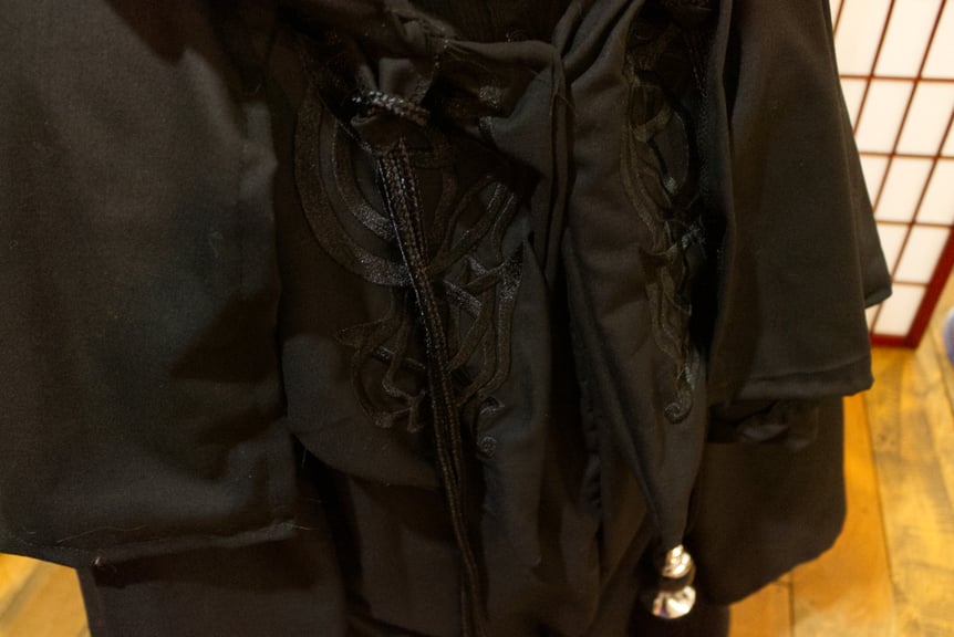 A close up of the swooping black-on black embroidered details of the robe. The robe is a soft close weave Italian linen in deep black, with highly textured and stylized sweeping embroidery.