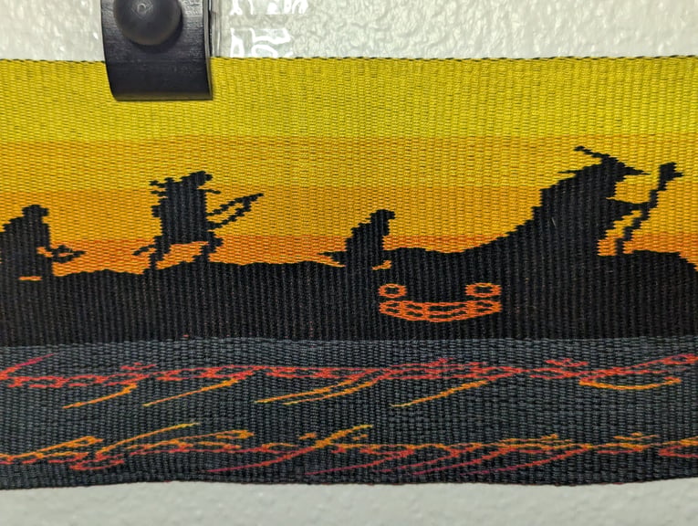 Fifth of six close ups of a tapestry depicting Gimli, Legolas, Frodo, Gandalf, and Harold the Creepy Doll below them and above part of the One Ring’s inscription.