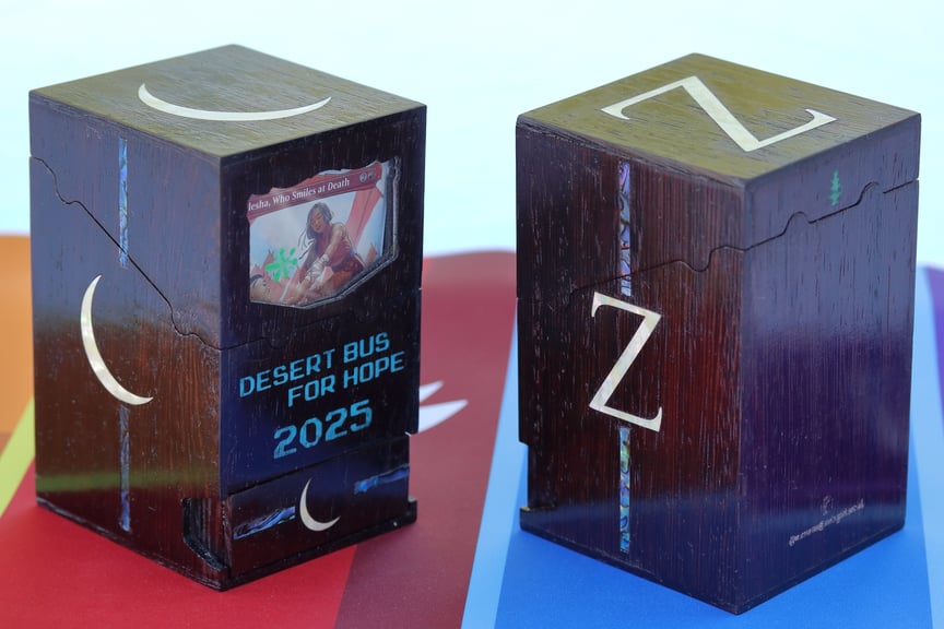 A closer view of the Night Watch and Zeta Shift boxes, still closed. The Night Watch box is seen from the front; the card visible through the window is Alesha, Who Smiles at Death. The Zeta Shift box is seen from behind, revealing a small green pine tree engraved in the back of the lid and a maker's mark at the bottom of the box. Blurry reflections on the boxes show the glossy finish.