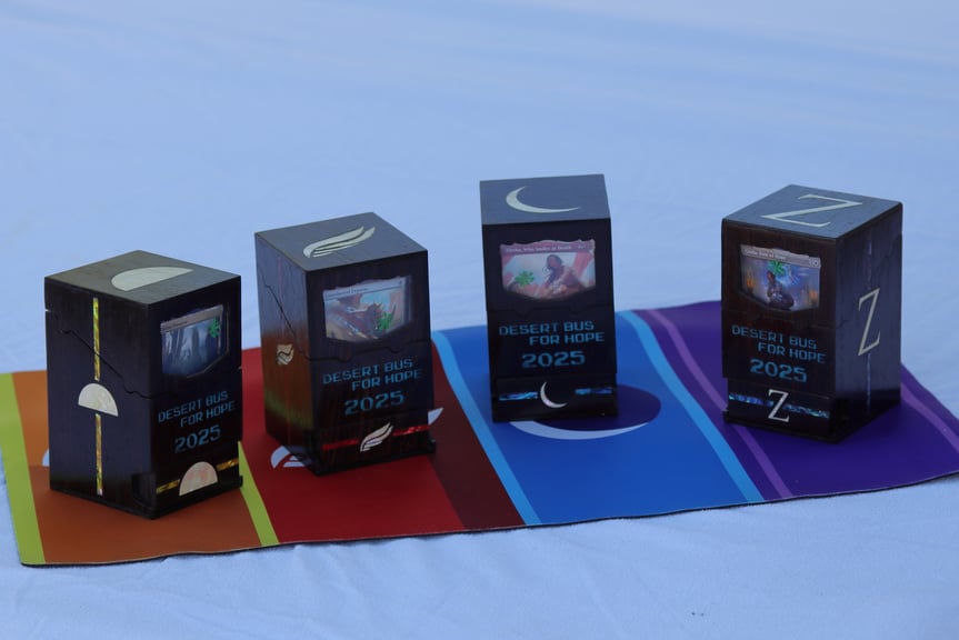 Four closed deck boxes, sitting on a Desert Bus playmat. The boxes are made from a dark brown hardwood. The sides and tops of the boxes each have the logo of a Desert Bus shift, and the sides also have a stripe in a color of the same shift. The fronts of the boxes have a clear window showing the cards within, and the words "Desert Bus For Hope 2025" inlaid in blue. The bottom of each box is a drawer, also with the shift logo and a colored stripe.