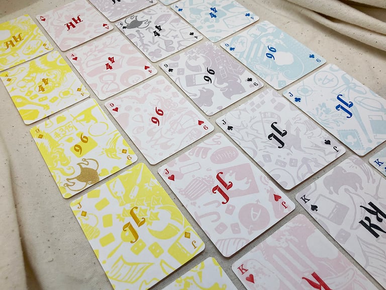 Several of the cards from each suit. The backdrop of each card's face is a pastel wallpaper of all the various joker designs, in a pale version of that suit's color; the pips and ranks of each card are printed on top in darker, more saturated colors, simply as letters and numbers, so as not to cover too much of the backdrop.