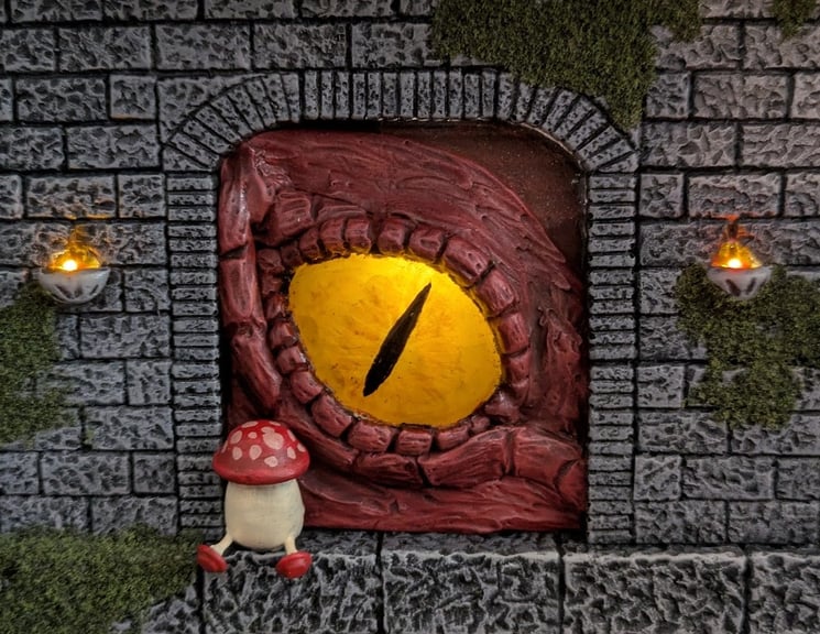 A close-up of the giant red dragon eye, inset into a large archway. A small walking mushroom sits in the arch in front of it. Both are flanked by two small flame sconces on the wall. The sconces and the eye are both lit with golden LEDs.