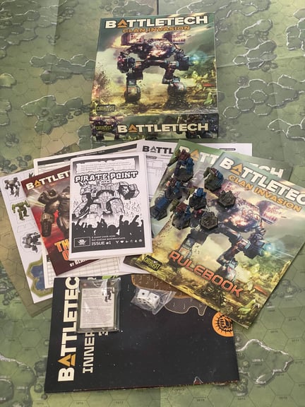 a spread of everything that comes in the prize. 5 Omnimech minis and 2 bases of Elementals and a truck, 2 double-sided maps, punchcard of additional mechs and terrain, rulebook, record sheets, Clan Invasion primer, novella, Pirate Point issue 1, Alpha Strike cards, reference sheet, 2 d6, and a map of the Inner Sphere

