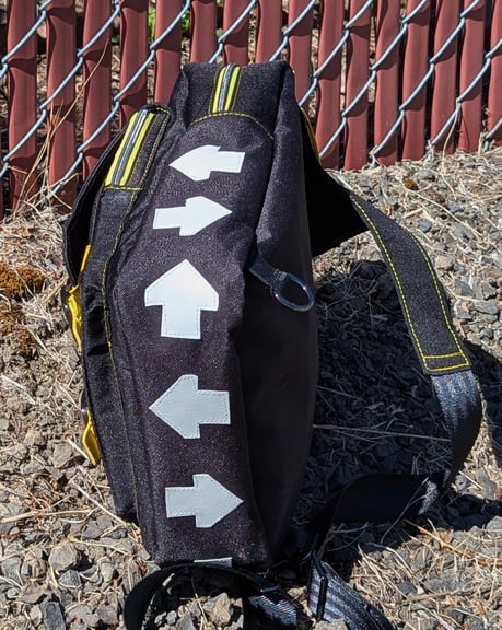 A black canvas backpack layed on it's side on gravel, showing the white vinyl arrows that wrap around the body of the bag, forming the Hellbomb strategem code.