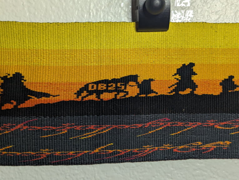 Second of six close ups of a tapestry depicting Aragorn, Sam leading Bill, Boromir, and Pippin above part of the One Ring’s inscription.