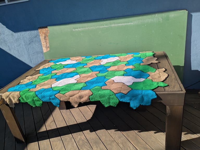 A knitted blanket outside on a rectangular, wooden table. The blanket is shorter than the table, but it's wide enough to hang over on both of the long edges. The blanket is made from 48 copies of a turtle shape, in sky blue, sand yellow, air-freshener green, and lane-line white. Around the outside and between turtles there are borders in asphalt black. The turtle shape was discovered in 2023 as an aperiodic monotile: it can tile the plane, but it can't form a repeating pattern. The white turtles are mirror images of the other turtles, and the turtles all appear in an assortment of rotations.
