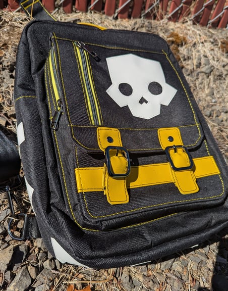 A black canvas backpack with yellow vinyl accents and a prominent white vinyl applique of the Helldivers 2 skull image, resting on gravel.