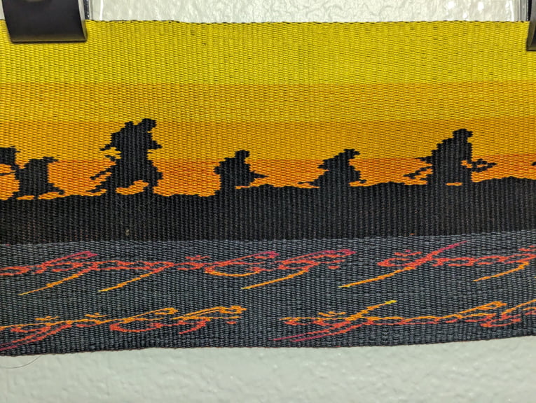 Third of six close ups of a tapestry depicting Sam, Boromir, Pippin, Merry, and Gimli above part of the One Ring’s inscription.