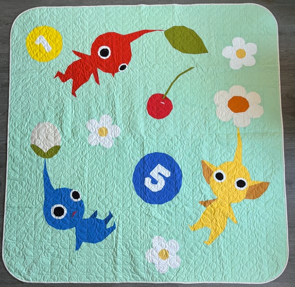 A flat lay of a mint green quilt with three Pikmin, red, yellow and blue along with some posy pellets, flowers and a cherry. 