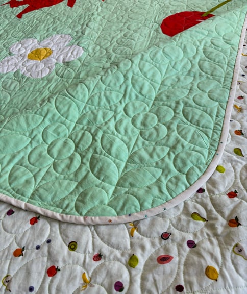 A detail shot of the quilting motif, daisies and leaves. As well as, showing off the rounded corners and the cute quilt white quilt backing that has various small fruits on it. 