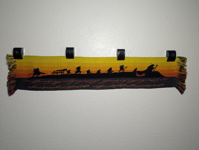 Hanging tablet woven tapestry of the Fellowship of the Ring’s silhouettes walking to Mordor in black & sunset colors.  Below is the Ring of Power’s inscription over two lines in fiery and smoky colors.