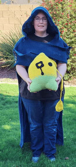 5'5" (165 cm) tall person from the front wearing a dark blue cape with hood holding a yellow and green orb fairy plush with 3 triangles trailing from it's head.  The cape is inspired from Legend of Zelda: Echoes of Wisdom and the plush is of the character Tri.