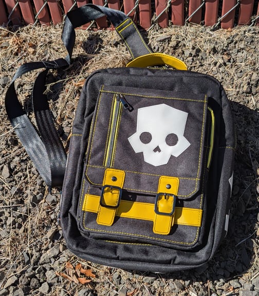 A black canvas backpack with yellow vinyl accents and a prominent white vinyl applique of the Helldivers 2 skull image, resting on gravel.