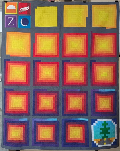 Quilt front. Each year of fundraising data is represented by a 13 x 13 piece block, with the individual pieces of the block spiraling out from the center of the block. Distance along the "spiral" represents run time in 1-hour increments and the colors of the pieces represent the total amount of money raised during the run. The two non-data blocks in the upper-left and lower-right corners are embroidered shift banners and a pixelated version of the Desert Bus for Hope logo, respectively.