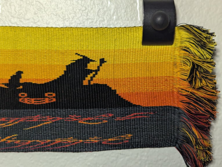 Sixth of six close ups of a tapestry depicting Frodo, Gandalf, and Harold the Creepy Doll below them and above part of the One Ring’s inscription.