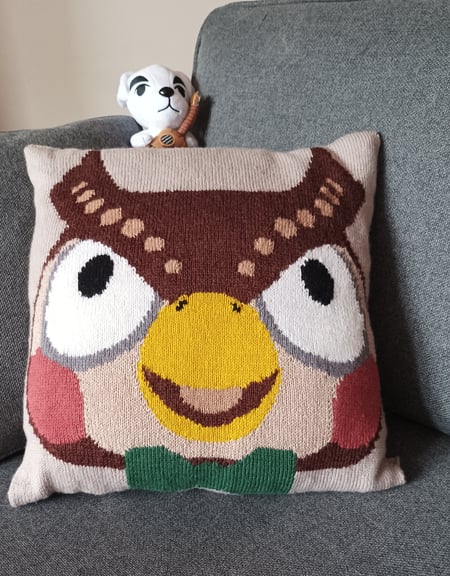 A photo of the Blathers cushion posed on a grey couch, with the head of a K.K. Slider plush poking out from behind it.